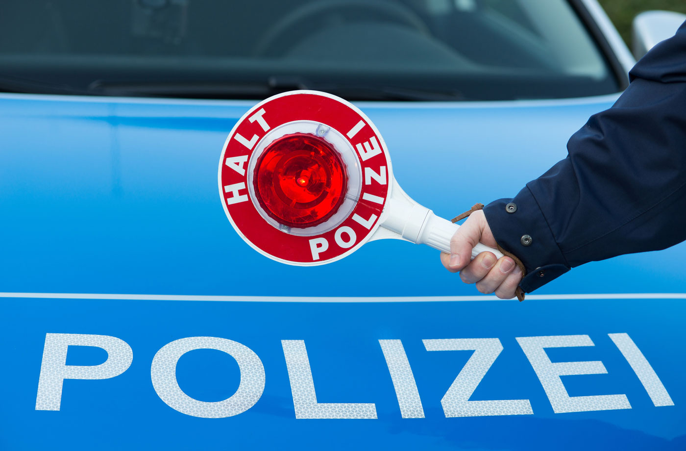 Arm der eine "HALT POLIZEI" Kelle hält mit Polizeischriftzug im Hintergrund